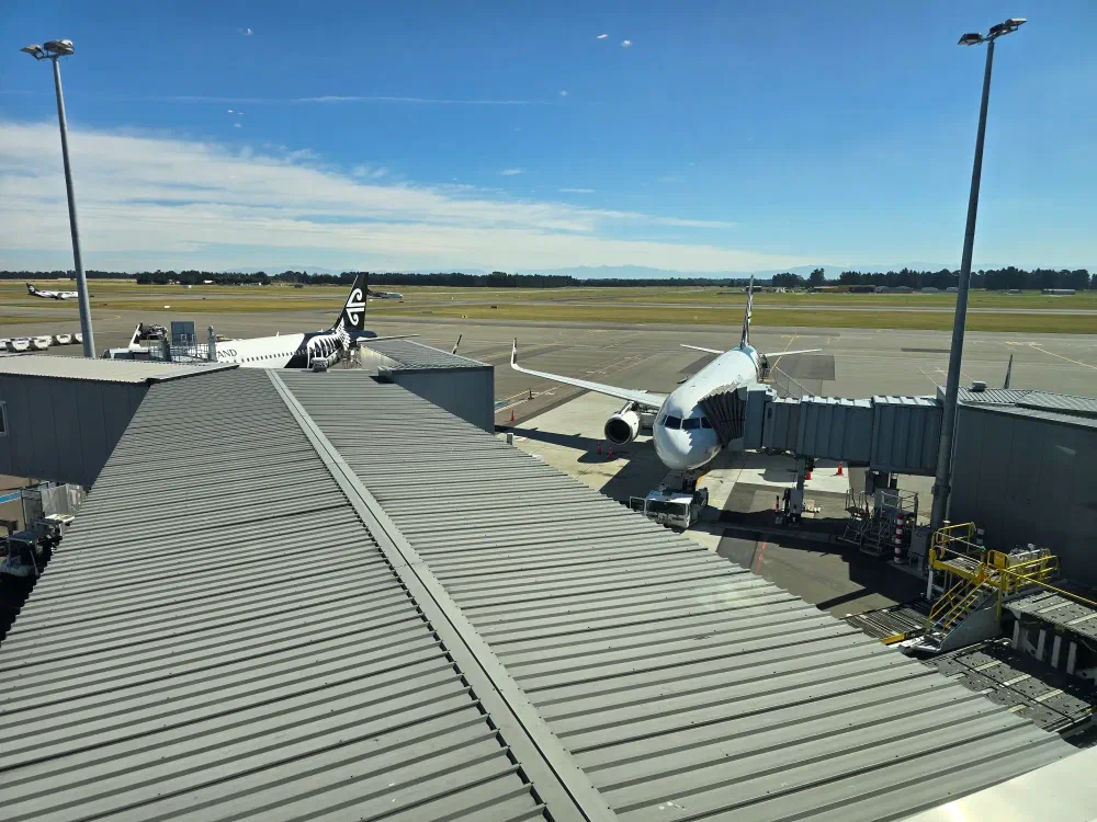 Overhead view of an Air New Zealand plane at a gate, attached to a jet bridge, with another plane visible in the distance on the tarmac. Christchurch Airport, New Zealand.