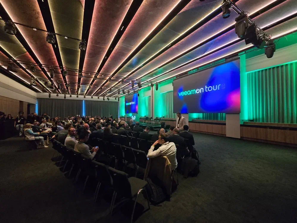 A wide shot of a modern conference hall during the VeeamOn Tour event in Melbourne. The room is filled with attendees seated in rows facing a stage, which has a large screen that reads &ldquo;VeeamOn Tour&rdquo;. The room is lit with colorful pink and green architectural lighting.