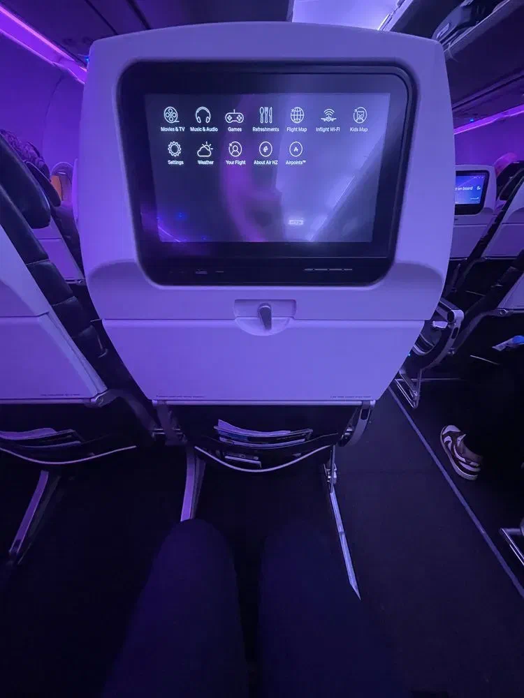 A view from an airplane seat looking forward at the in-flight entertainment screen. The dark cabin is illuminated by purple mood lighting.