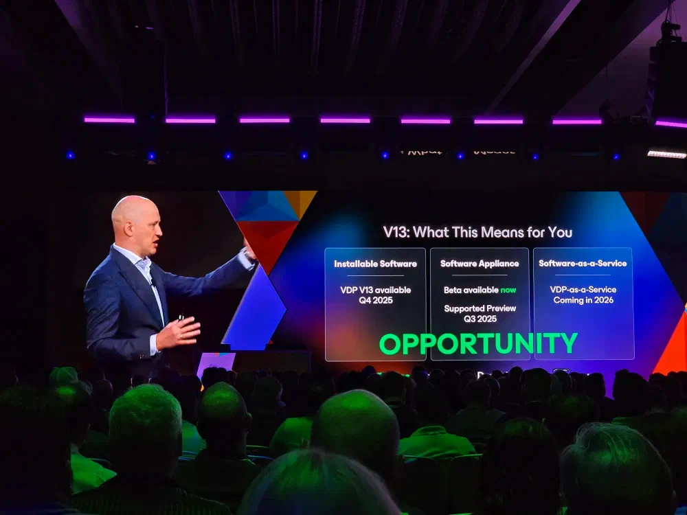 A speaker on stage at a conference points to a large screen displaying “V13: What This Means for You” and details about software availability and “OPPORTUNITY”. An audience is visible in the foreground.