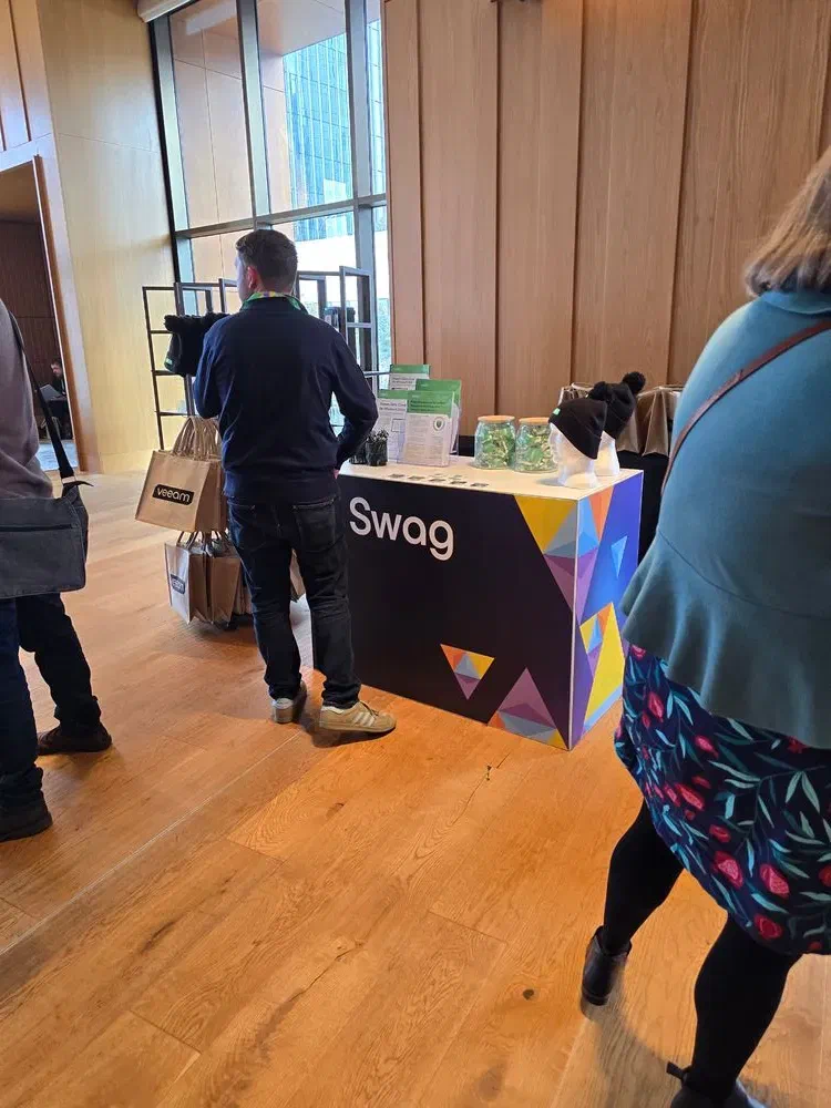 An event attendee stands at the VeeamOn Tour &ldquo;Swag&rdquo; station, which is decorated with colorful geometric patterns. Veeam-branded tote bags and beanies are visible on the counter.