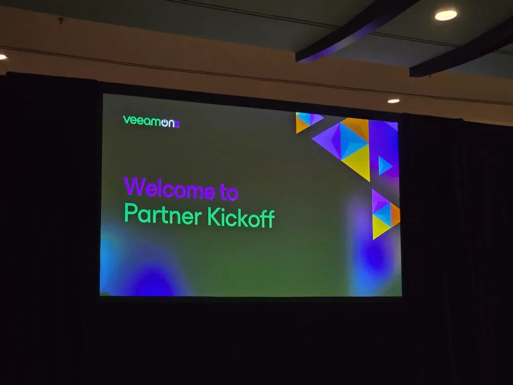 A screen displays “veeamon Welcome to Partner Kickoff” with colorful abstract shapes in the background.