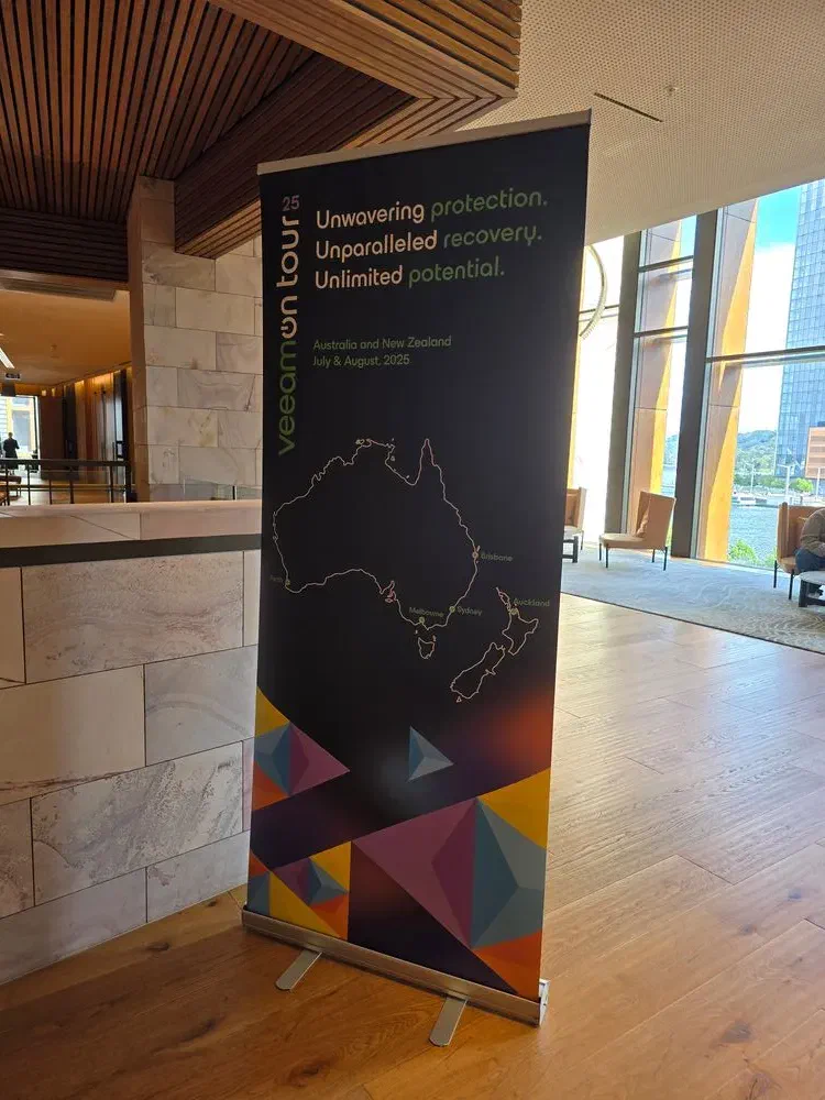  A tall, black VeeamOn Tour 2025 pull-up banner standing in a modern building lobby. The banner reads &ldquo;Unwavering protection. Unparalleled recovery. Unlimited potential.&rdquo; and shows a map of Australia and New Zealand.