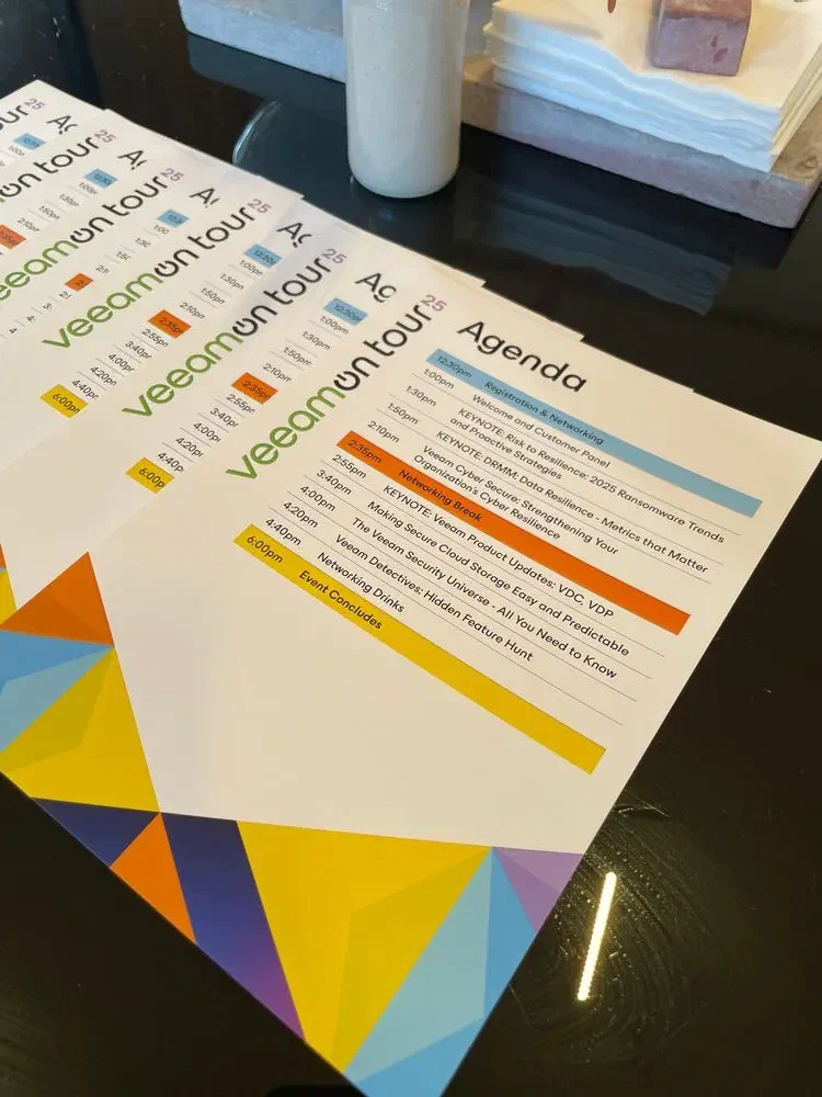 A close-up of several printed VeeamOn Tour agendas fanned out on a glossy black table. The agenda lists session times and topics for the event, with a white vase in the background.